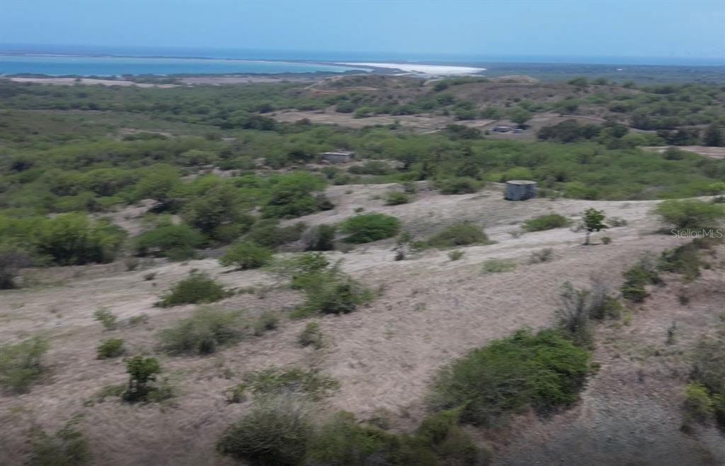 An aerial view shows a grassy, shrub-covered landscape with a small concrete structure near the coast — Km 12.1 PR 303, CABO ROJO, PR