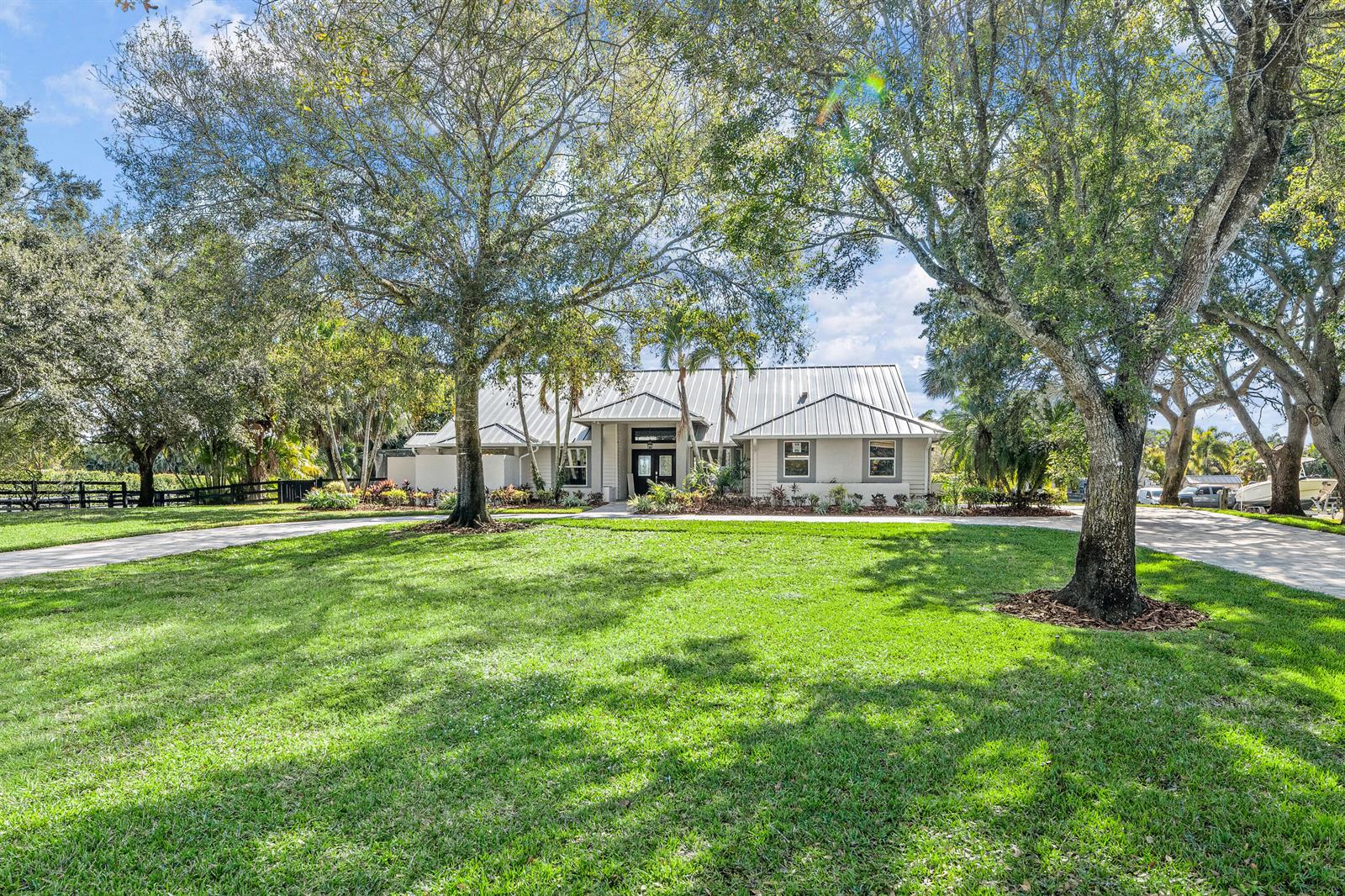 A single-story house with a metal roof sits behind a large lawn and mature trees — 14564 Rolling Rock Place, Wellington FL 33414, WELLINGTON, FL