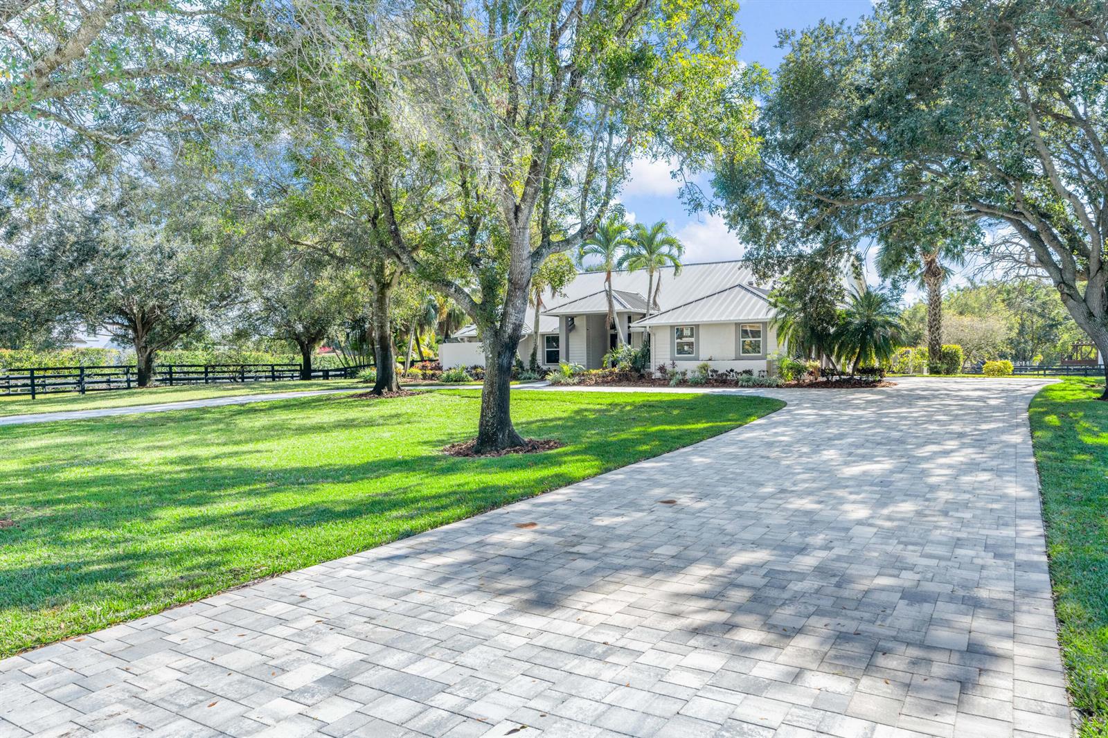 A paved driveway leads to a light-colored house surrounded by large trees and green lawns — 14564 Rolling Rock Place, Wellington FL 33414, WELLINGTON, FL