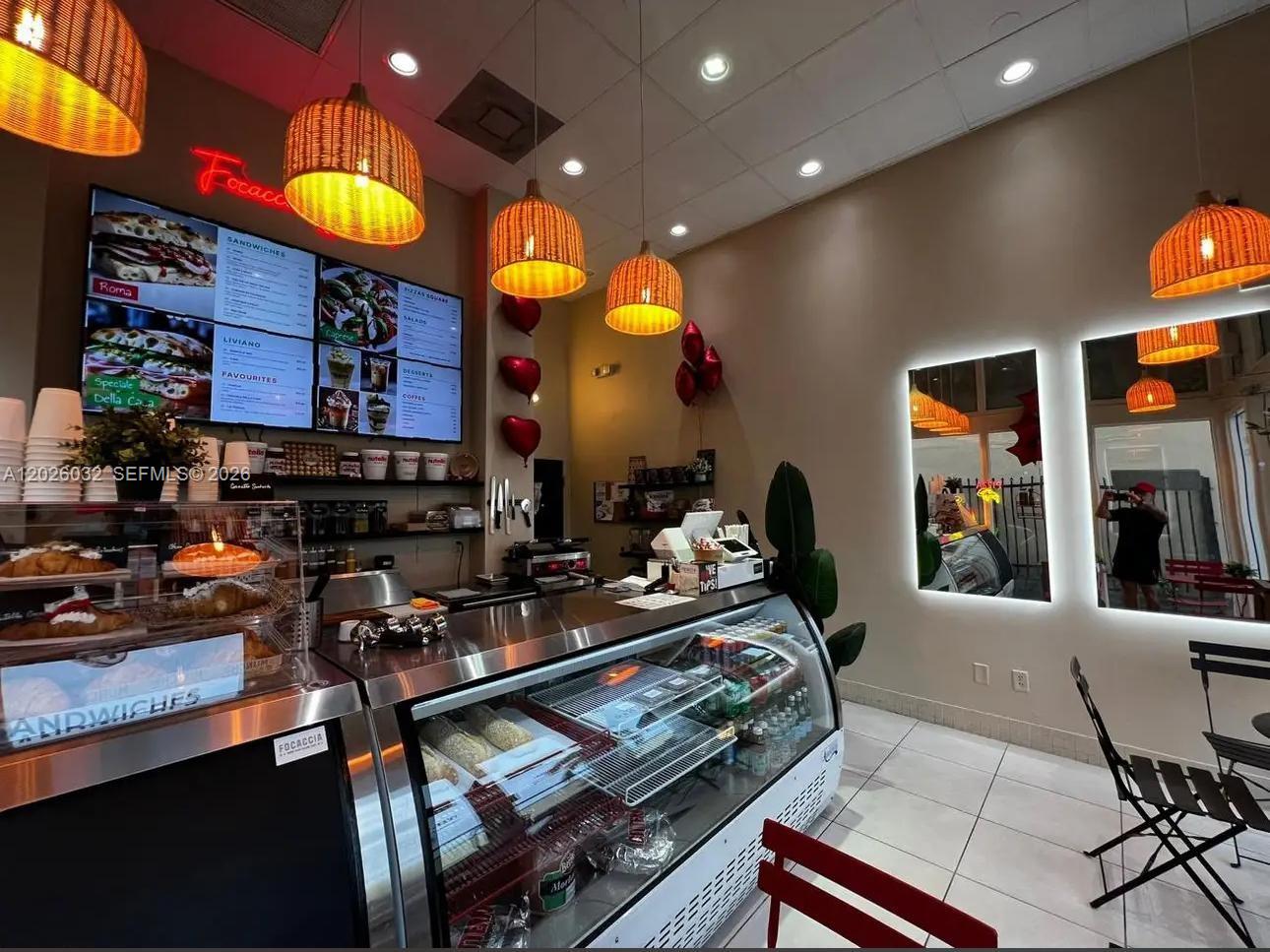 A brightly lit cafe interior features a service counter, display cases, and woven pendant lights — NORTH MIAMI BEACH, FL