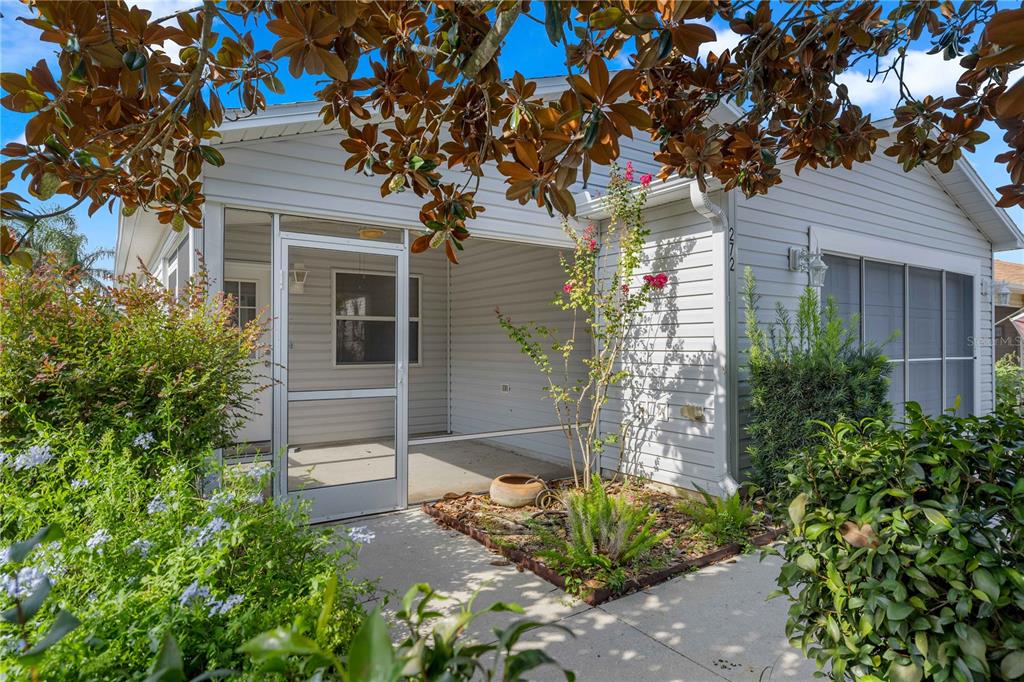 A screened-in porch with light siding and a concrete walkway sits beside a small garden — 272 EMMALEE PLACE, THE VILLAGES, FL