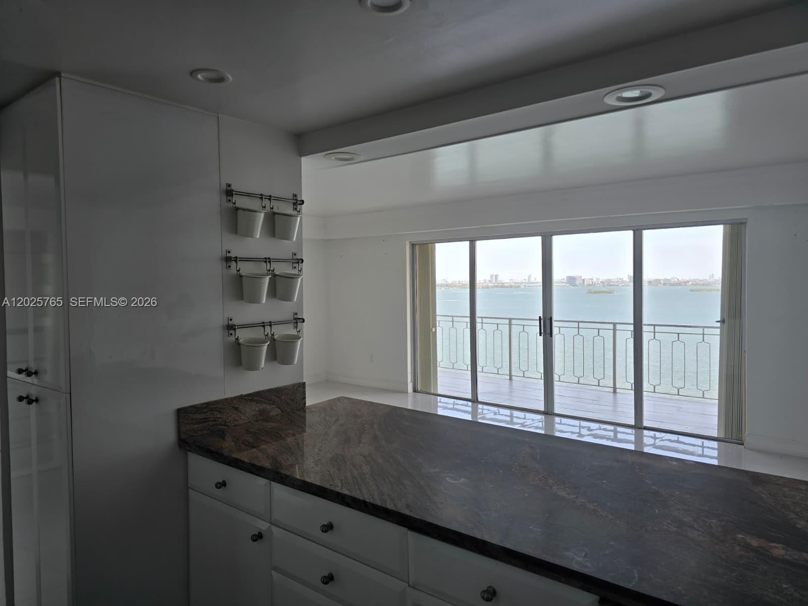 A kitchen with white cabinets and dark granite countertops overlooks a balcony and water view — 11111 Biscayne Blvd # 17D, Miami FL 33181, MIAMI, FL