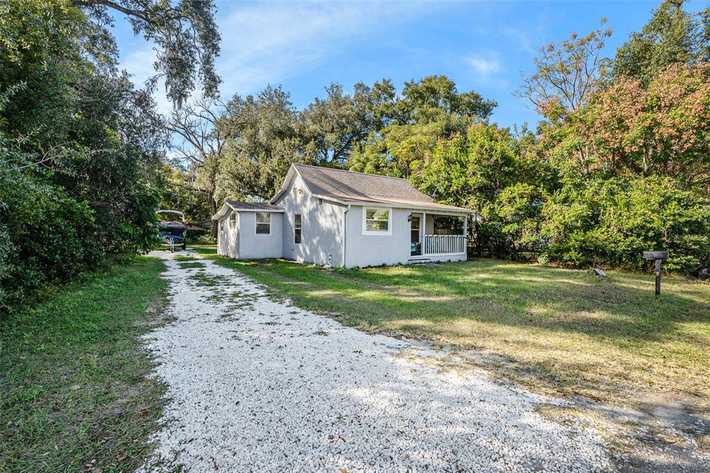 A light-colored house with a gravel driveway sits on a wooded lot — 4921 6TH STREET, ZEPHYRHILLS, FL