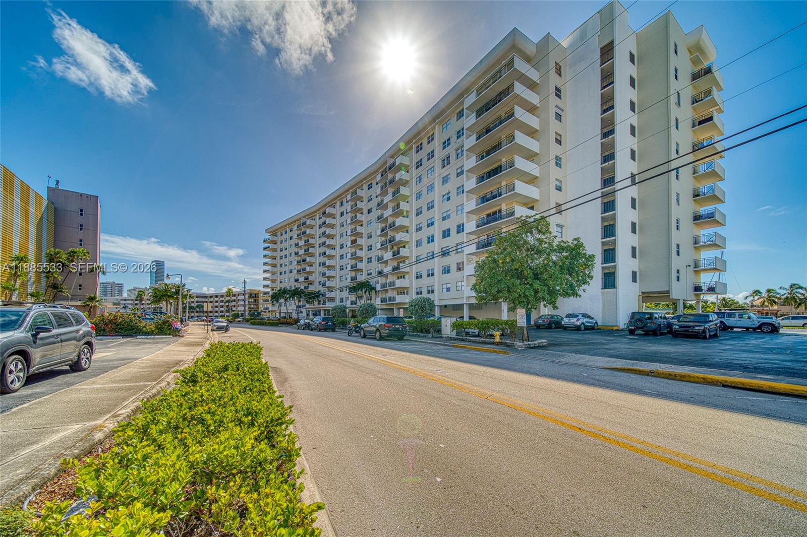 A multi-story apartment building stands along a paved street with parked cars and landscaping — 401 Golden Isles Dr # 908, Hallandale Beach FL 33009, HALLANDALE BEACH, FL