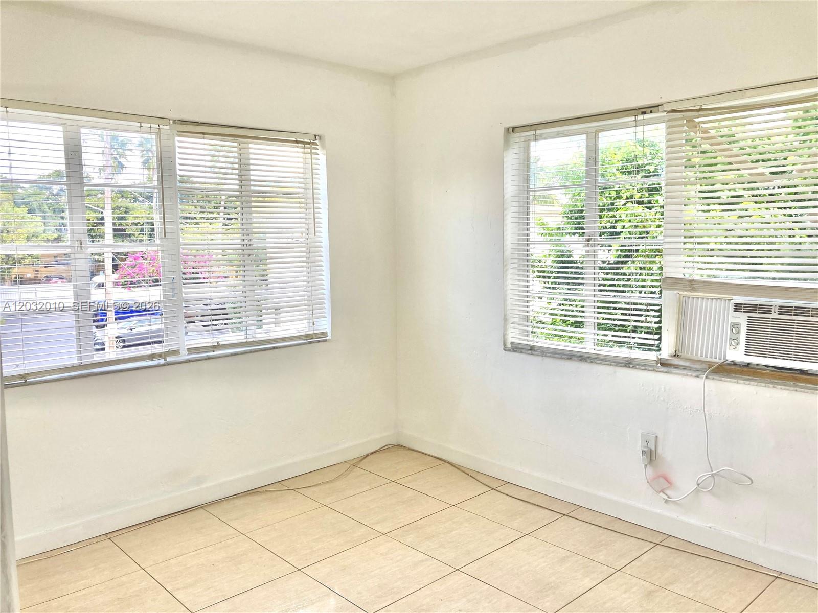 An empty room features white walls, beige tile flooring, two windows with blinds, and an air conditioner — 625 83rd St # 44, Miami Beach FL 33141, MIAMI BEACH, FL