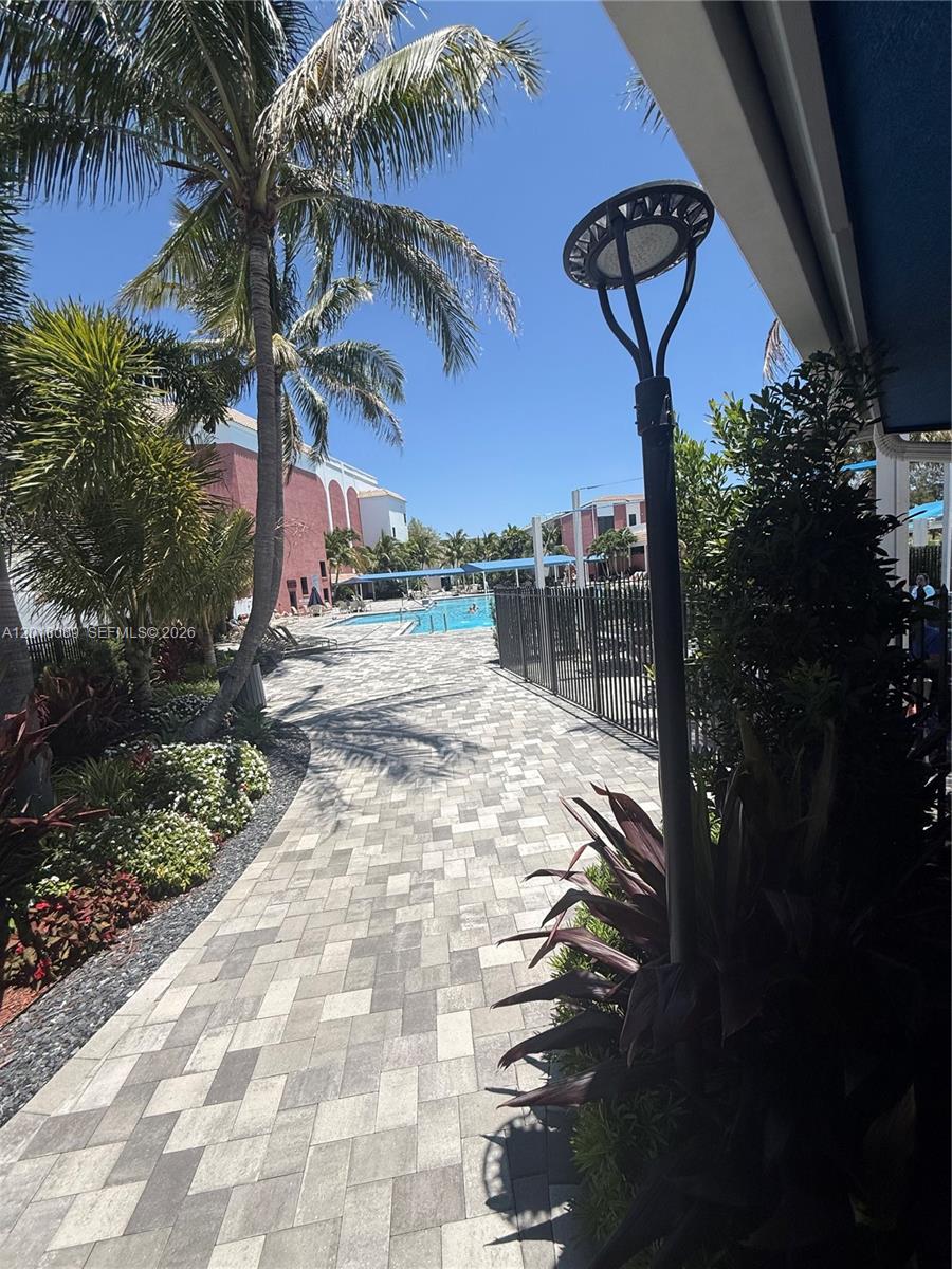 A paved walkway leads toward a swimming pool surrounded by palm trees and building structures — 4047 Lyndhurst K # 4047, Deerfield Beach FL 33442, DEERFIELD BEACH, FL