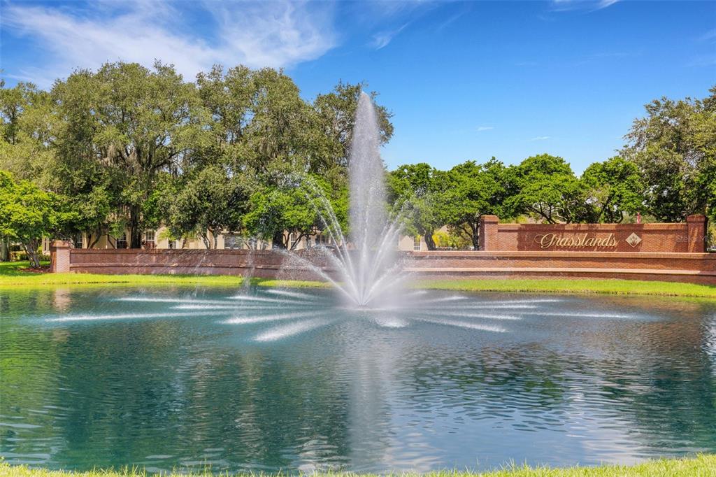 A large fountain sprays water into a pond in front of a brick community sign — 516 GRASSLANDS VILLAGE CIRCLE, LAKELAND, FL