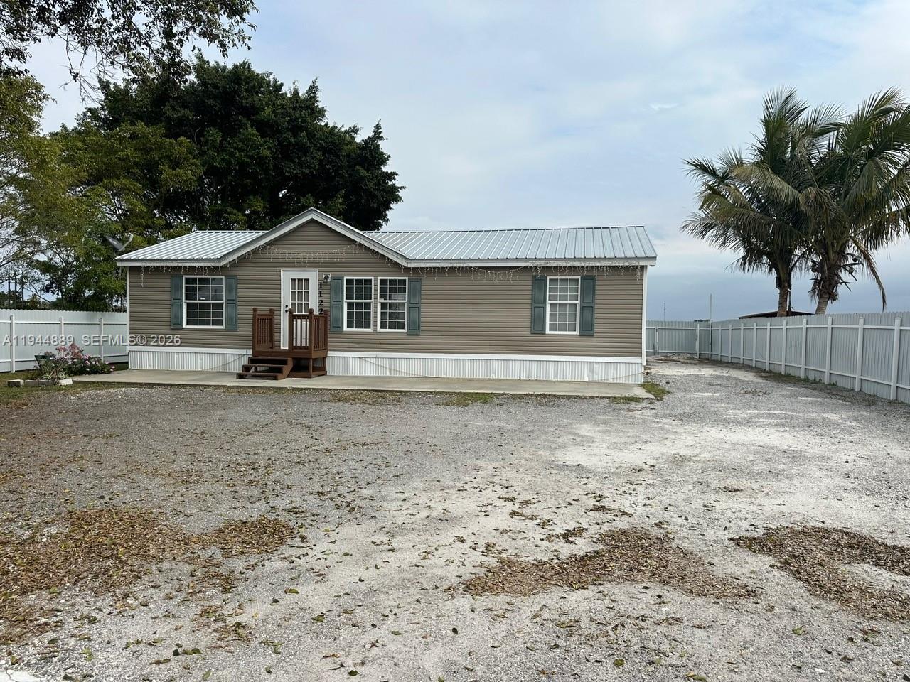 A single-story manufactured home with tan siding and a metal roof sits on a gravel lot — 1122 JOHNSON RD, Clewiston FL 33440, CLEWISTON, FL