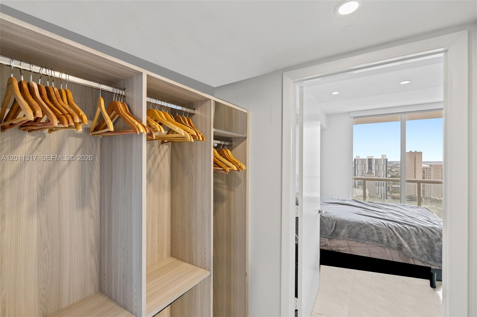 A walk-in closet with light wood shelving and hanging rods overlooks a bedroom with city views — 50 Biscayne Blvd # 4405, Miami FL 33132, MIAMI, FL