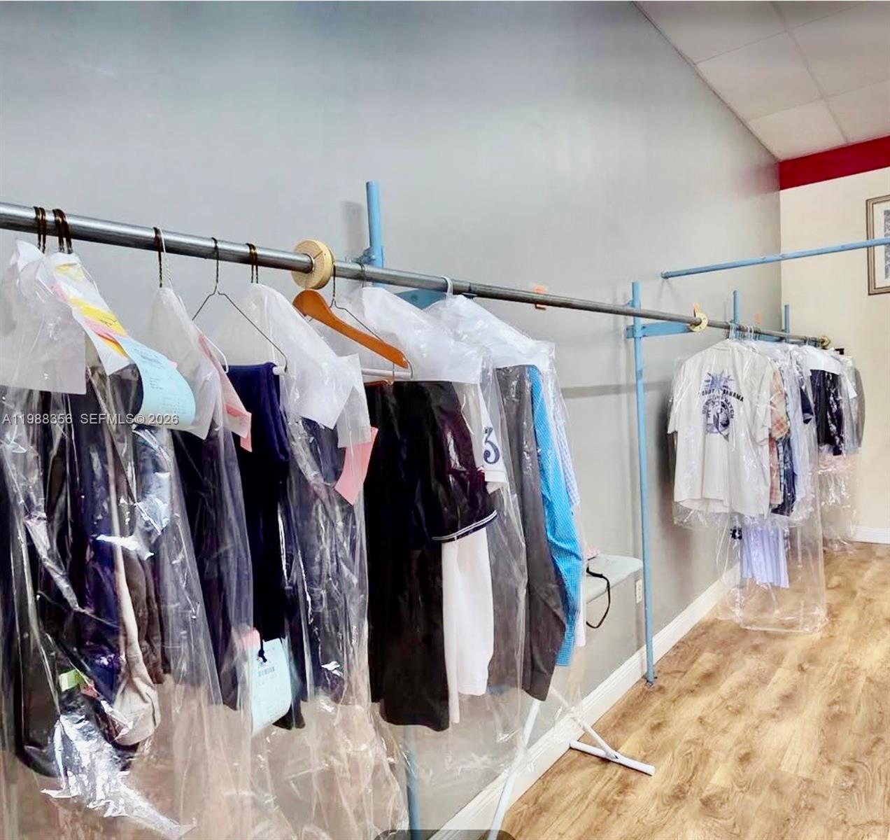 A room features a long metal clothing rack holding garments covered in plastic dry-cleaning bags — 4222 W 16th Ave, Hialeah FL 33012, HIALEAH, FL