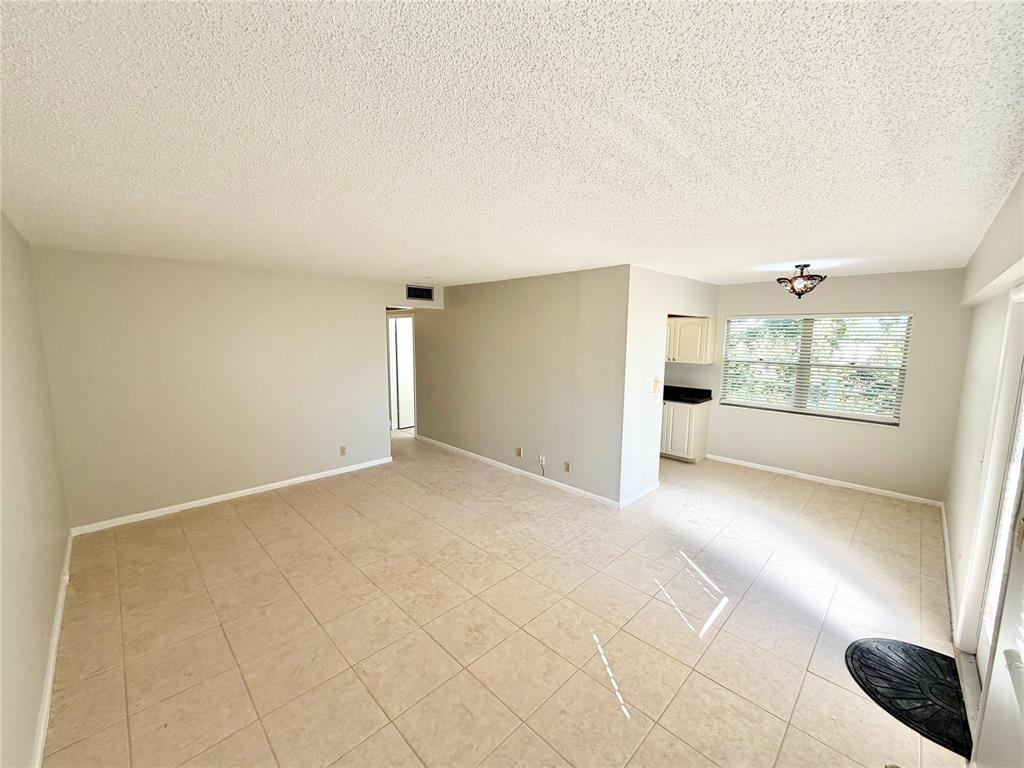 An empty living room with beige tile flooring, light-colored walls, and a small kitchen area — 84 Mansfield B # 84, Boca Raton FL 33434, BOCA RATON, FL