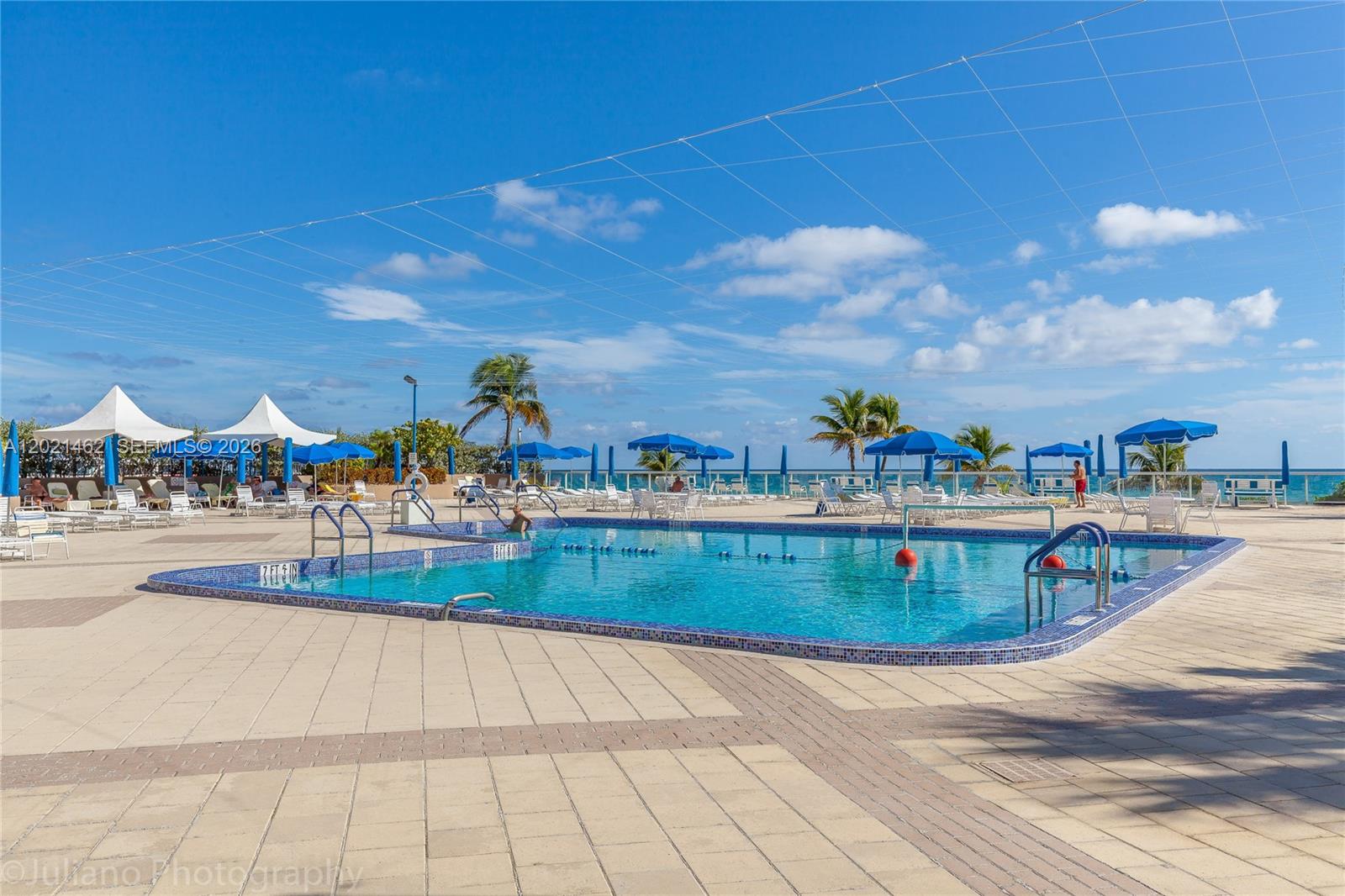 An outdoor swimming pool with blue tiles is surrounded by a paved patio and umbrellas — 2030 S Ocean Dr # 908, Hallandale Beach FL 33009, HALLANDALE BEACH, FL