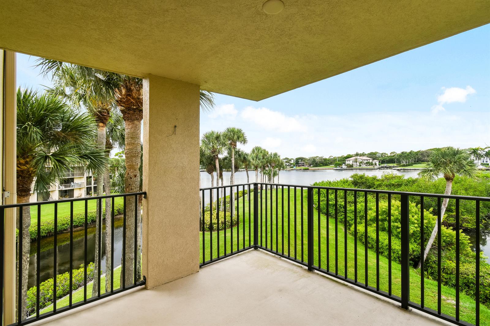 A covered balcony with black railings overlooks a grassy lawn, palm trees, and a waterway — 1701 Marina Isle Way # 301, Jupiter FL 33477, JUPITER, FL