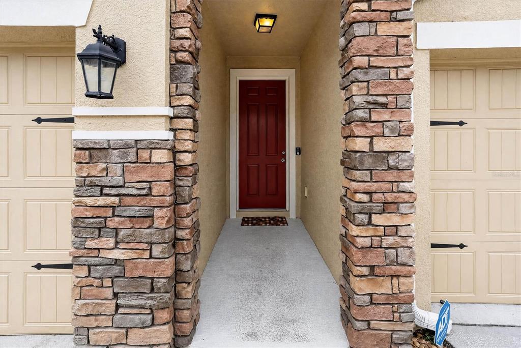 A red front door is centered between two stone-clad pillars and beige garage doors — 1608 MOHEGAN BOULEVARD, KISSIMMEE, FL