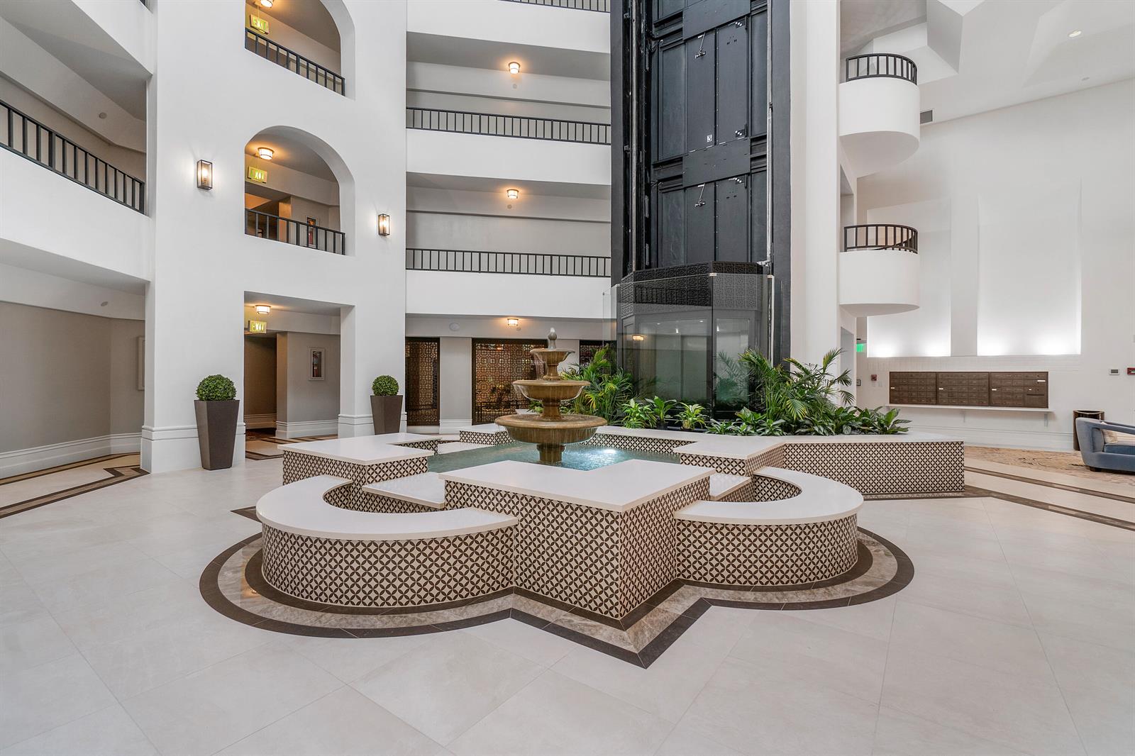 A multi-story atrium features a central tiered fountain surrounded by curved seating and balconies — 100 SE 5th Avenue # 410, Boca Raton FL 33432, BOCA RATON, FL