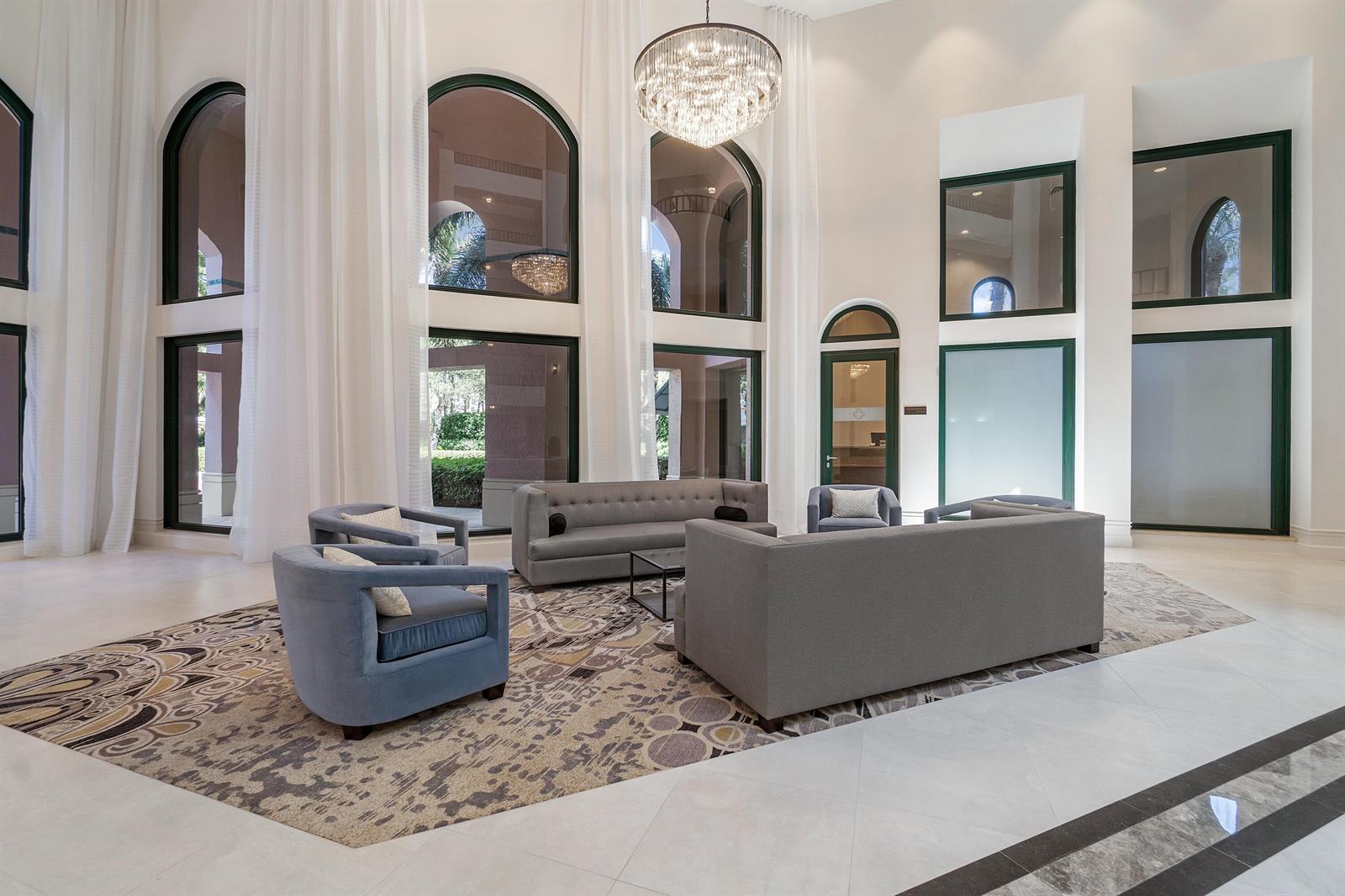 A spacious lobby features high ceilings, a crystal chandelier, grey furniture, and large arched windows — 100 SE 5th Avenue # 410, Boca Raton FL 33432, BOCA RATON, FL