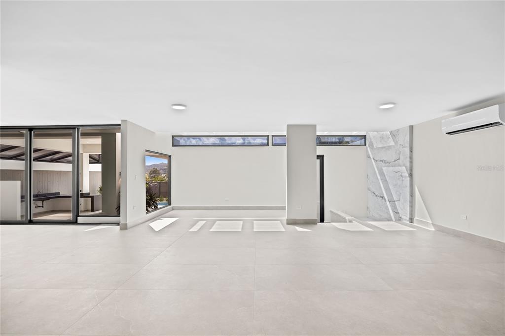 A bright, minimalist living space features large light-colored floor tiles, a marble accent wall, and sliding glass doors — La Villa de Torrimar REY JORGE 373, GUAYNABO, PR