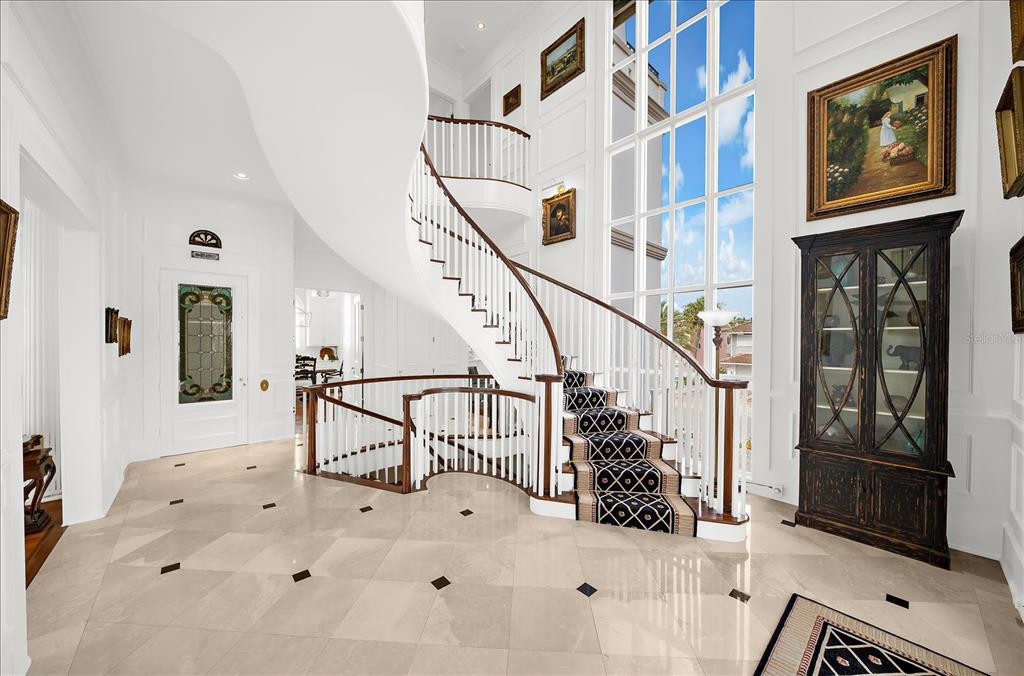 A grand foyer features a curved staircase with a patterned runner and large floor-to-ceiling windows — 810 ELDORADO AVENUE, CLEARWATER BEACH, FL