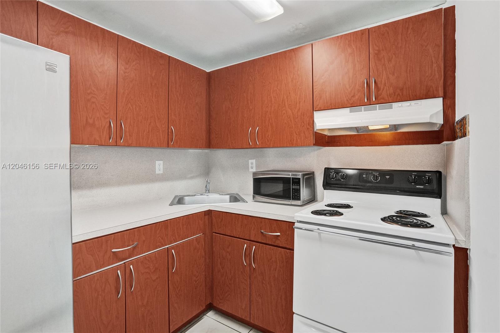 A small kitchen features wood-grain cabinets, white appliances, and a corner sink — 606 W 81st St # 323, Hialeah FL 33014, HIALEAH, FL