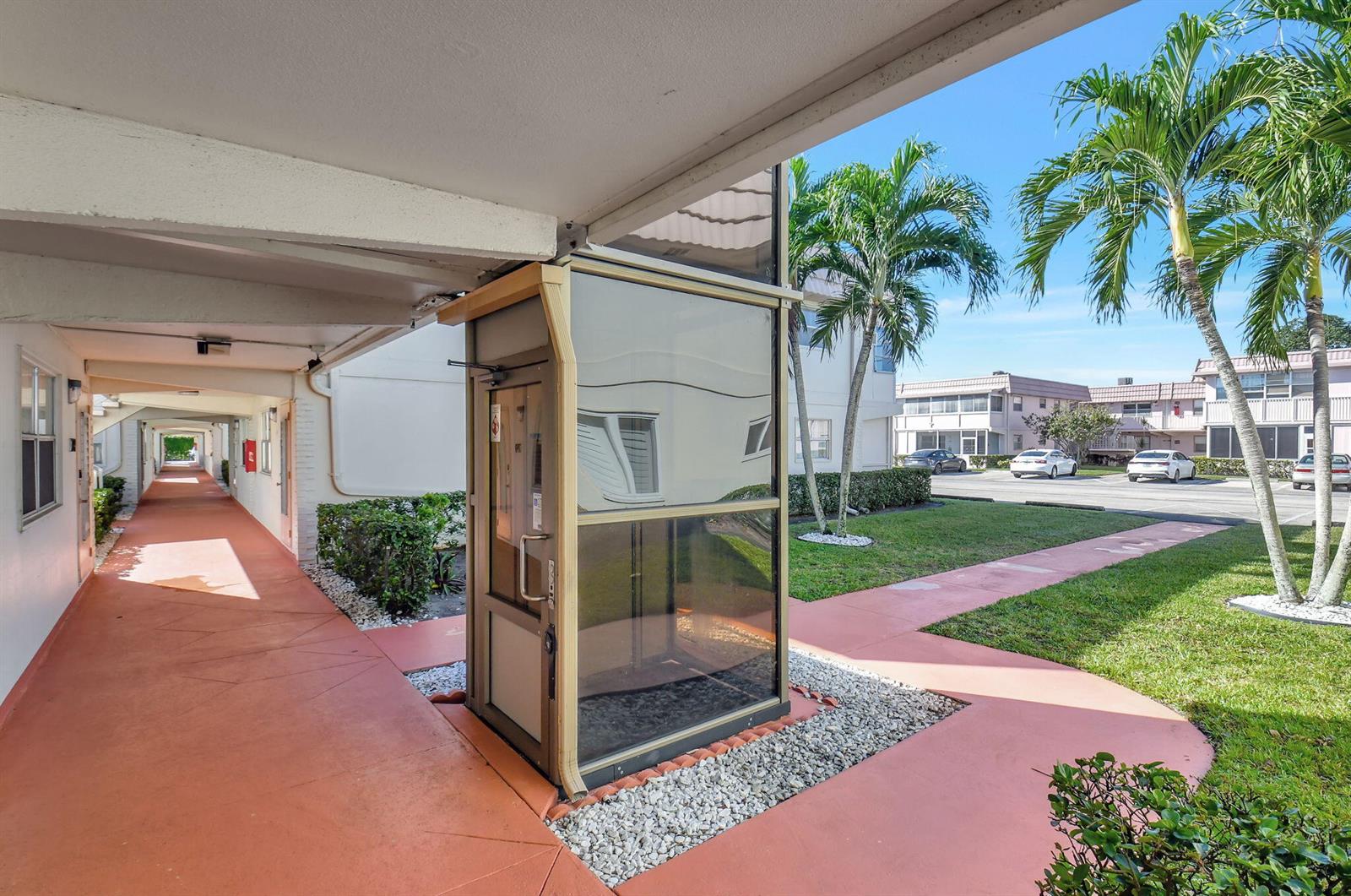 An outdoor elevator enclosure stands on a walkway next to a building and palm trees — 325 Saxony G, Delray Beach FL 33446, DELRAY BEACH, FL