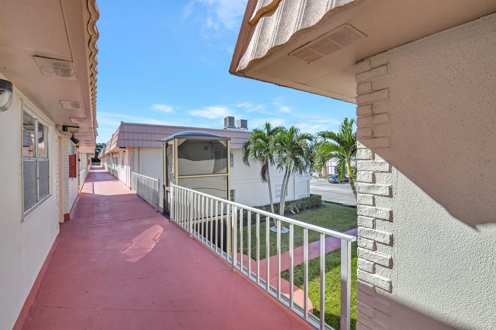 An outdoor walkway with a red floor and a metal lift overlooks a grassy courtyard — 325 Saxony G, Delray Beach FL 33446, DELRAY BEACH, FL