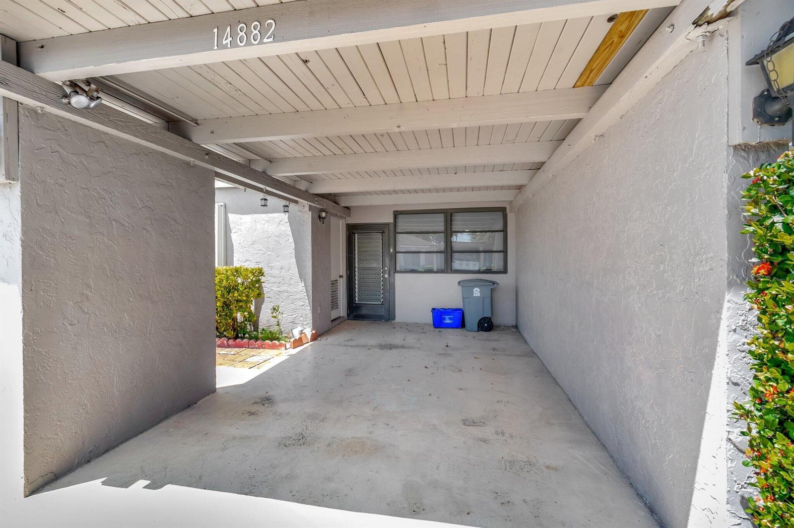 A covered concrete carport features a grey exterior wall, a front door, and a window — 14882 Wildflower Lane, Delray Beach FL 33446, DELRAY BEACH, FL