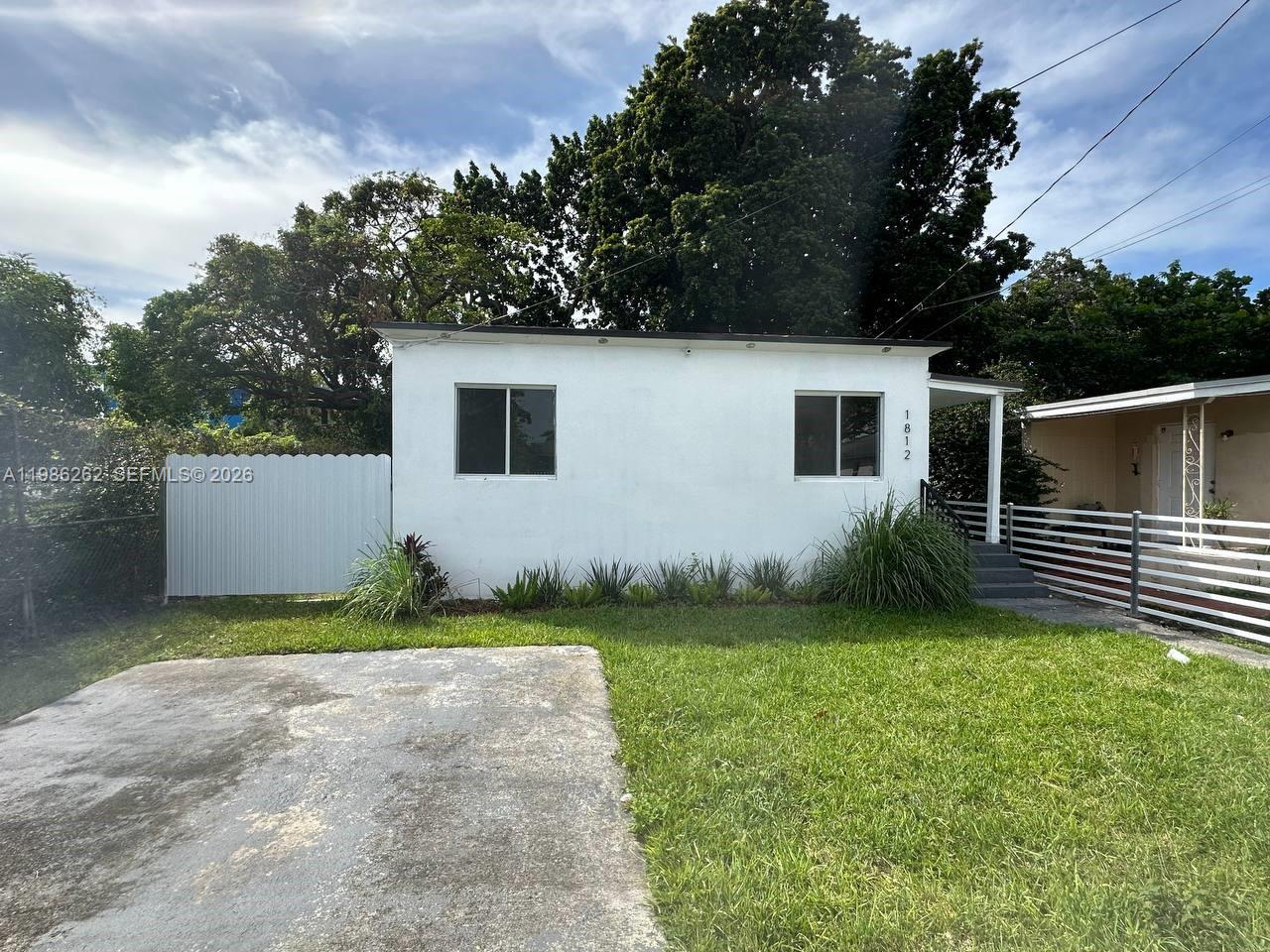 A small, white, single-story house features a concrete driveway and a white metal fence — 1812 NW 64th St, Miami FL 33147, MIAMI, FL