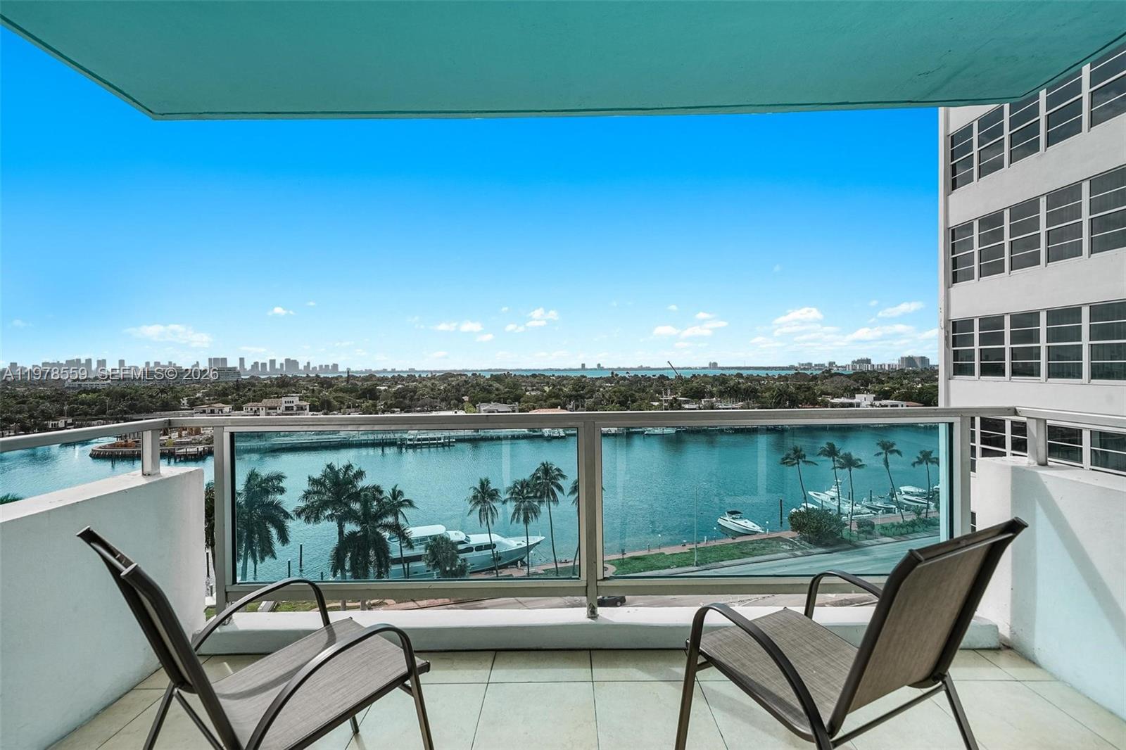 A balcony with two chairs overlooks a waterway, palm trees, and a distant city skyline — 5101 Collins Ave # 11T, Miami Beach FL 33140, MIAMI BEACH, FL