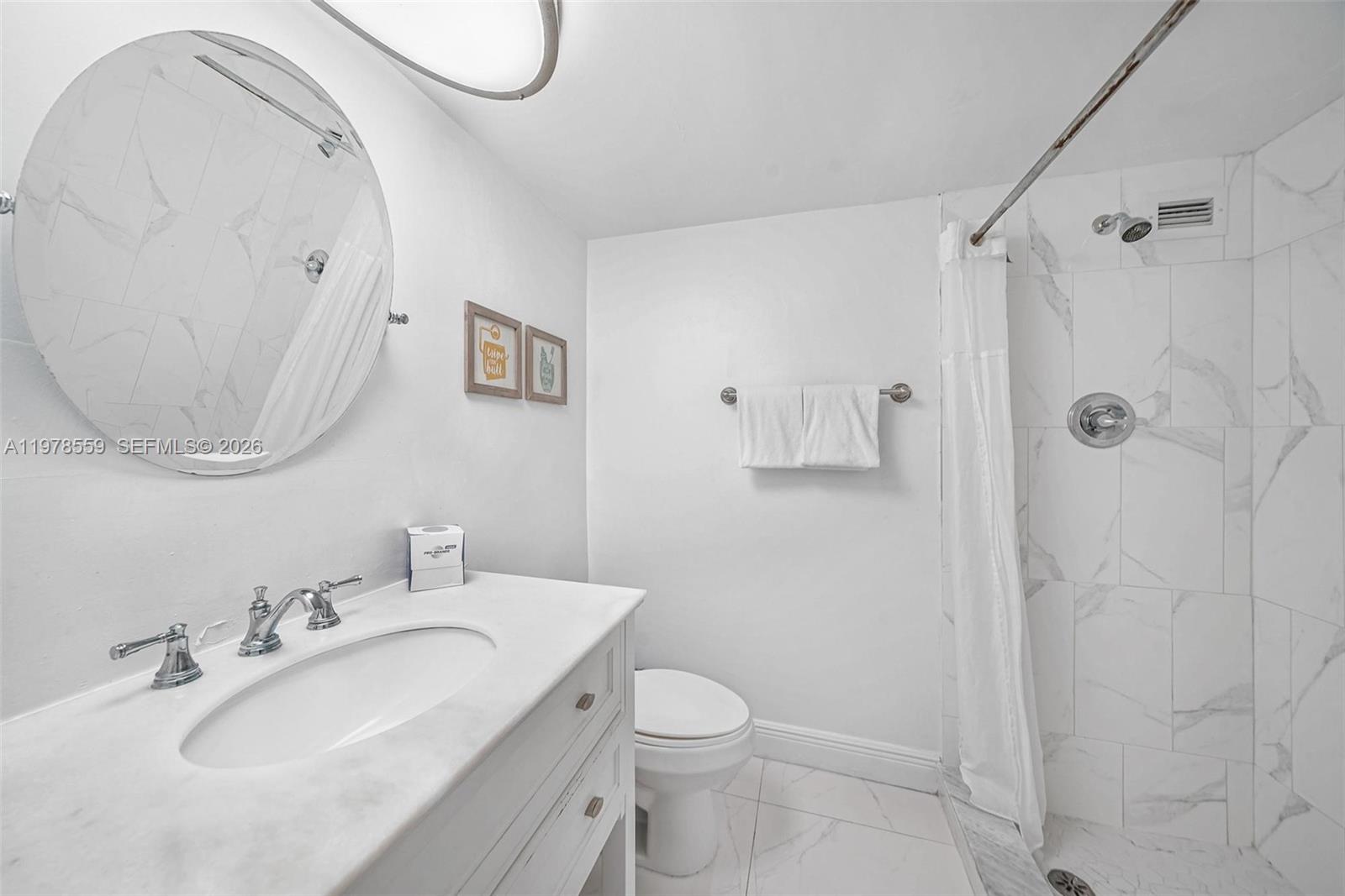 A white bathroom features a marble-topped vanity, a round mirror, and a tiled shower stall — 5101 Collins Ave # 11T, Miami Beach FL 33140, MIAMI BEACH, FL