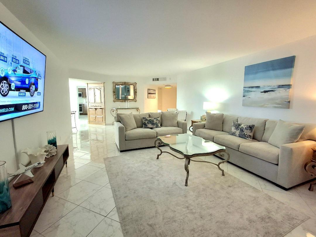 A living room features light-colored sofas, a glass coffee table, and a large wall-mounted television — 1340 S Ocean Boulevard # 1808, Pompano Beach FL 33062, POMPANO BEACH, FL