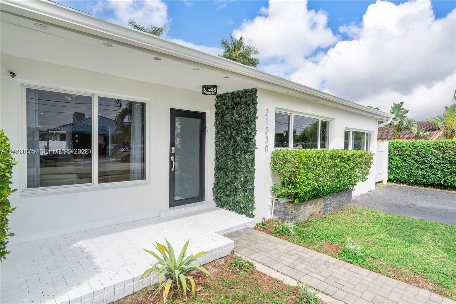 The exterior of a white house features a front porch, a green wall, and landscaping — 21230 NE 24th Ct # 0, Miami FL 33180, MIAMI, FL