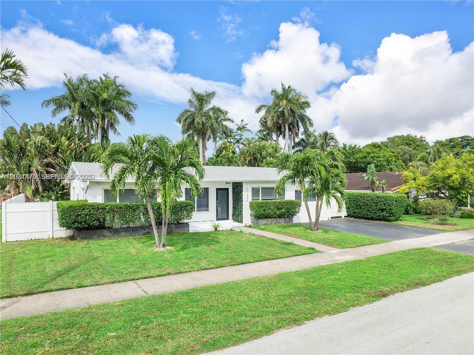 A single-story white house with a lawn, palm trees, and a driveway under a blue sky — 21230 NE 24th Ct # 0, Miami FL 33180, MIAMI, FL