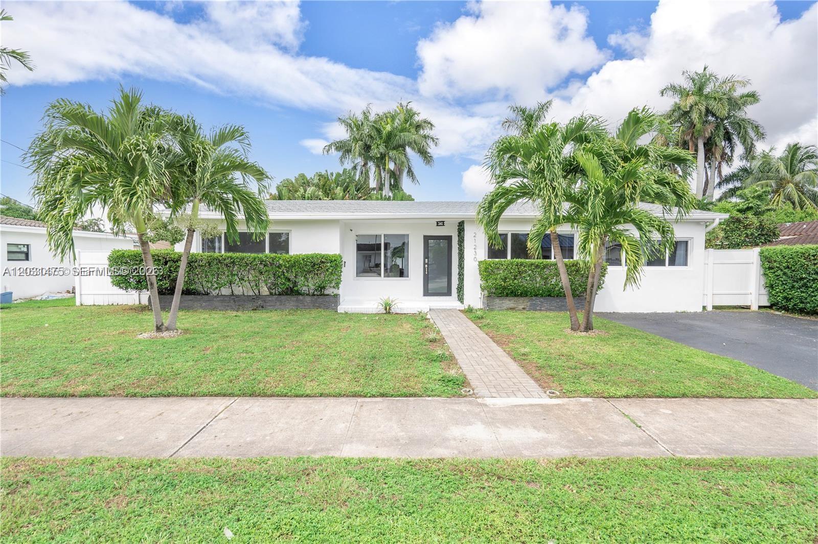 A white single-story house features a paved walkway, two palm trees, and manicured hedges — 21230 NE 24th Ct # 0, Miami FL 33180, MIAMI, FL