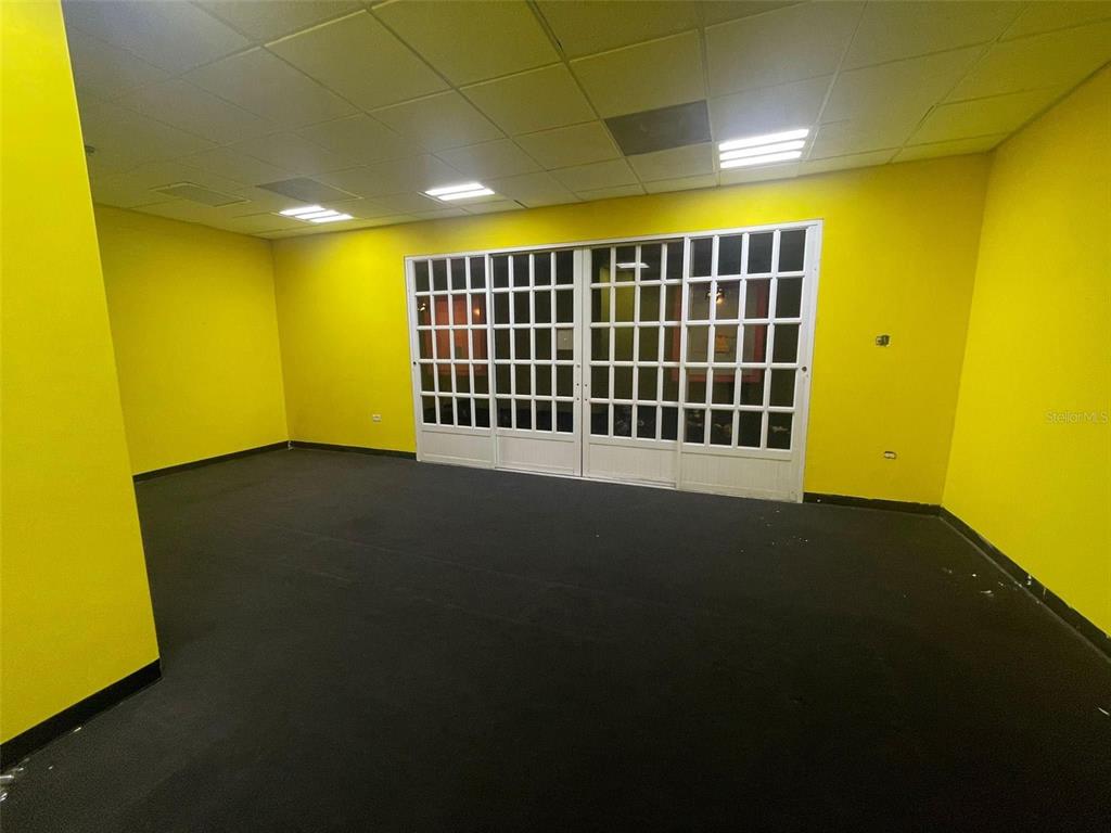 An empty room with bright yellow walls, black carpeting, and a white grid-paneled sliding door — 1 C. ISABEL MUÑOZ RIVERA, PONCE, PR
