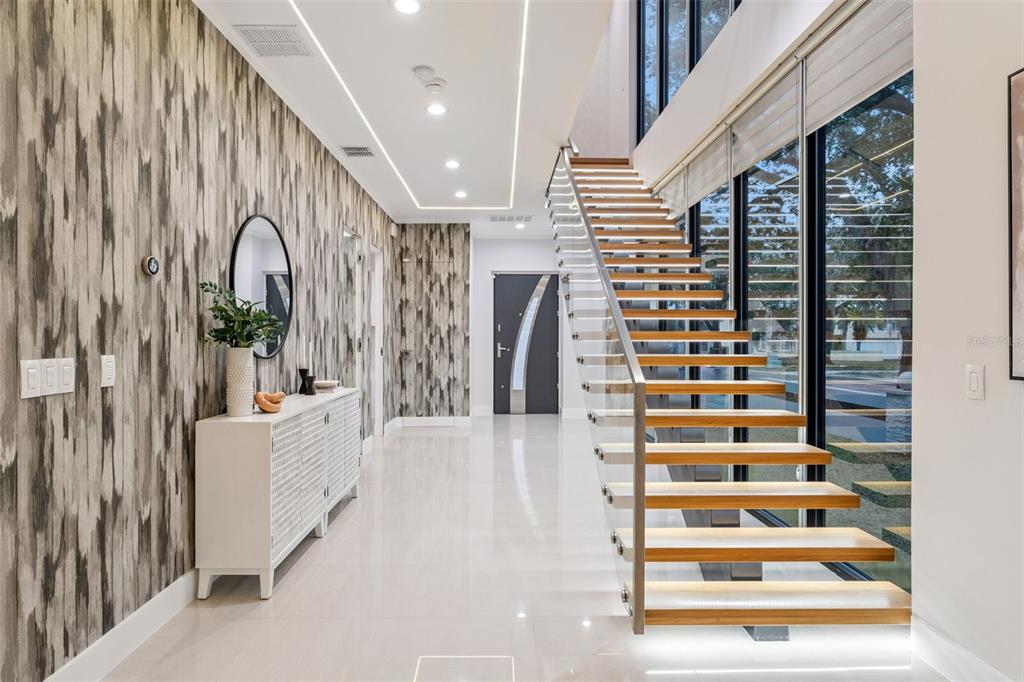 A modern entryway features a floating wooden staircase, glass railings, patterned wallpaper, and white flooring — 1903 N PARK AVENUE, WINTER PARK, FL