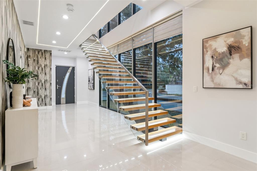 A modern entryway features a floating wooden staircase with glass railings and polished white floors — 1903 N PARK AVENUE, WINTER PARK, FL