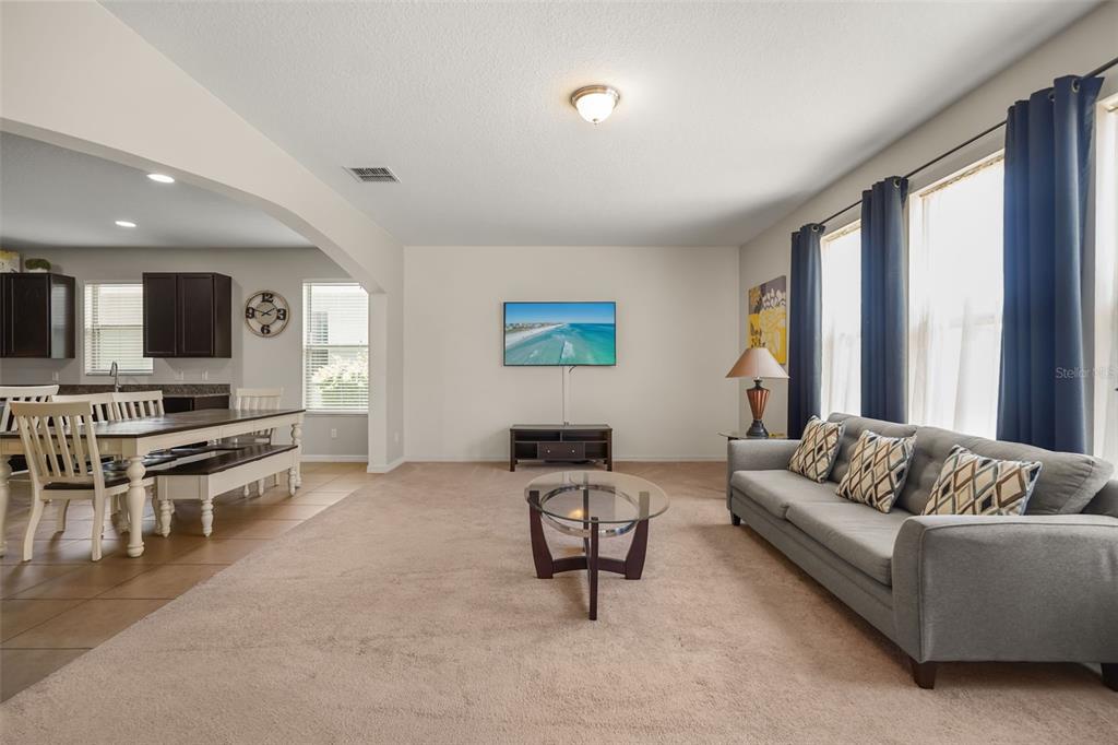 A living room with beige carpet, a grey sofa, a wall-mounted television, and an adjacent dining area — 1612 PINE MARSH LOOP, ST CLOUD, FL