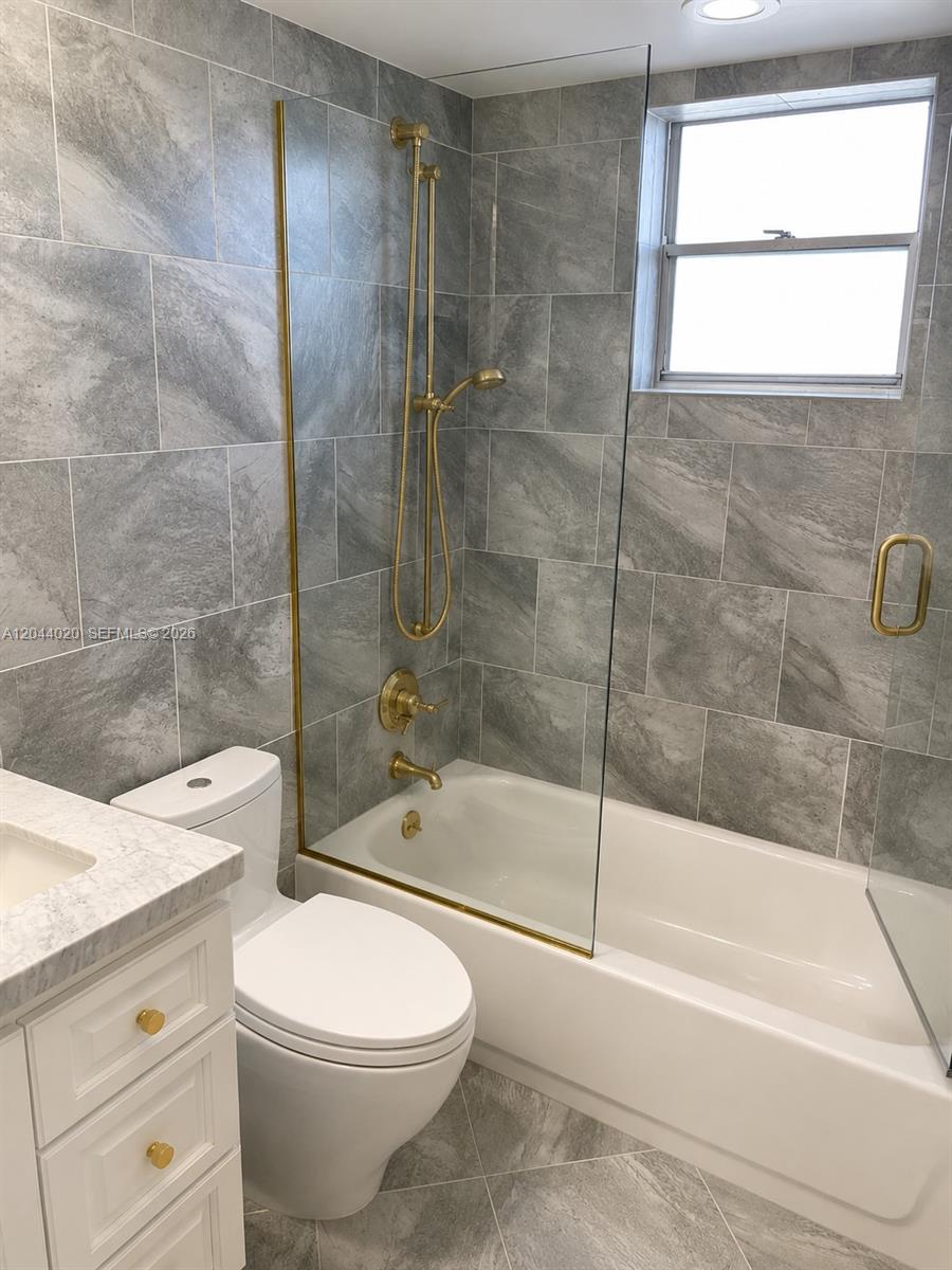 A bathroom features gray tile walls, a white bathtub, a glass panel, and gold fixtures — 219 NE 14th Ave # 306, Hallandale Beach FL 33009, HALLANDALE BEACH, FL