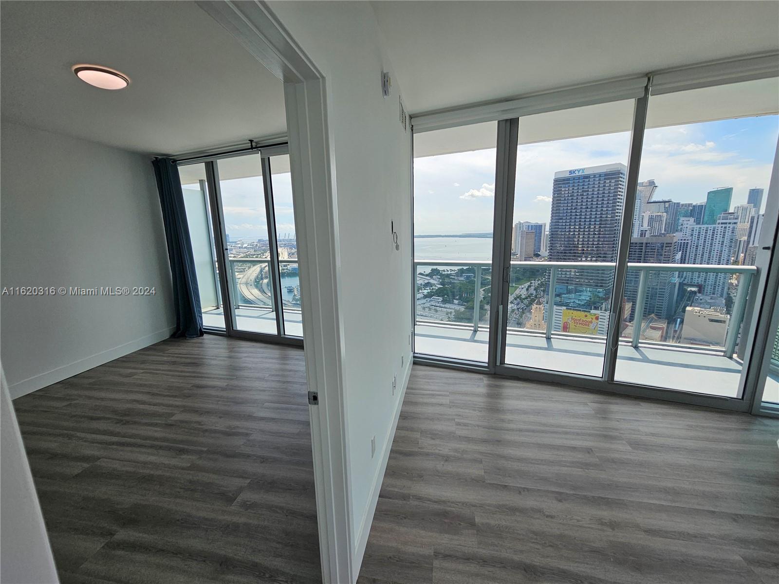An empty room with wood-look flooring and floor-to-ceiling windows overlooking a city skyline and water — 888 Biscayne Blvd # 2701, Miami FL 33132, MIAMI, FL