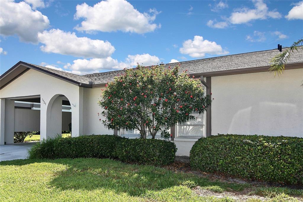 A single-story house exterior features a white stucco facade, a covered entryway, and landscaping — 1527 STEWART DRIVE UNIT 115, SARASOTA, FL