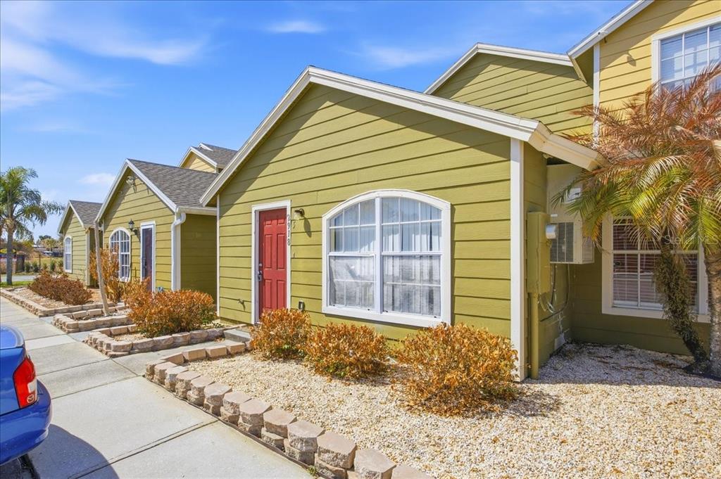 A row of olive-green townhomes with red front doors and stone-bordered landscaping under a blue sky — 2918 FAN PALM DRIVE Front unit, DAVENPORT, FL