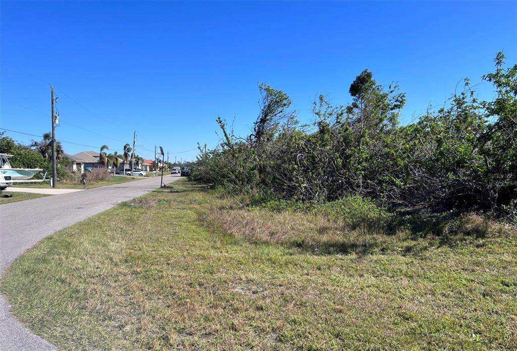 A paved road runs alongside a grassy lot with dense vegetation under a clear sky — 15531 AQUA CIRCLE, PORT CHARLOTTE, FL