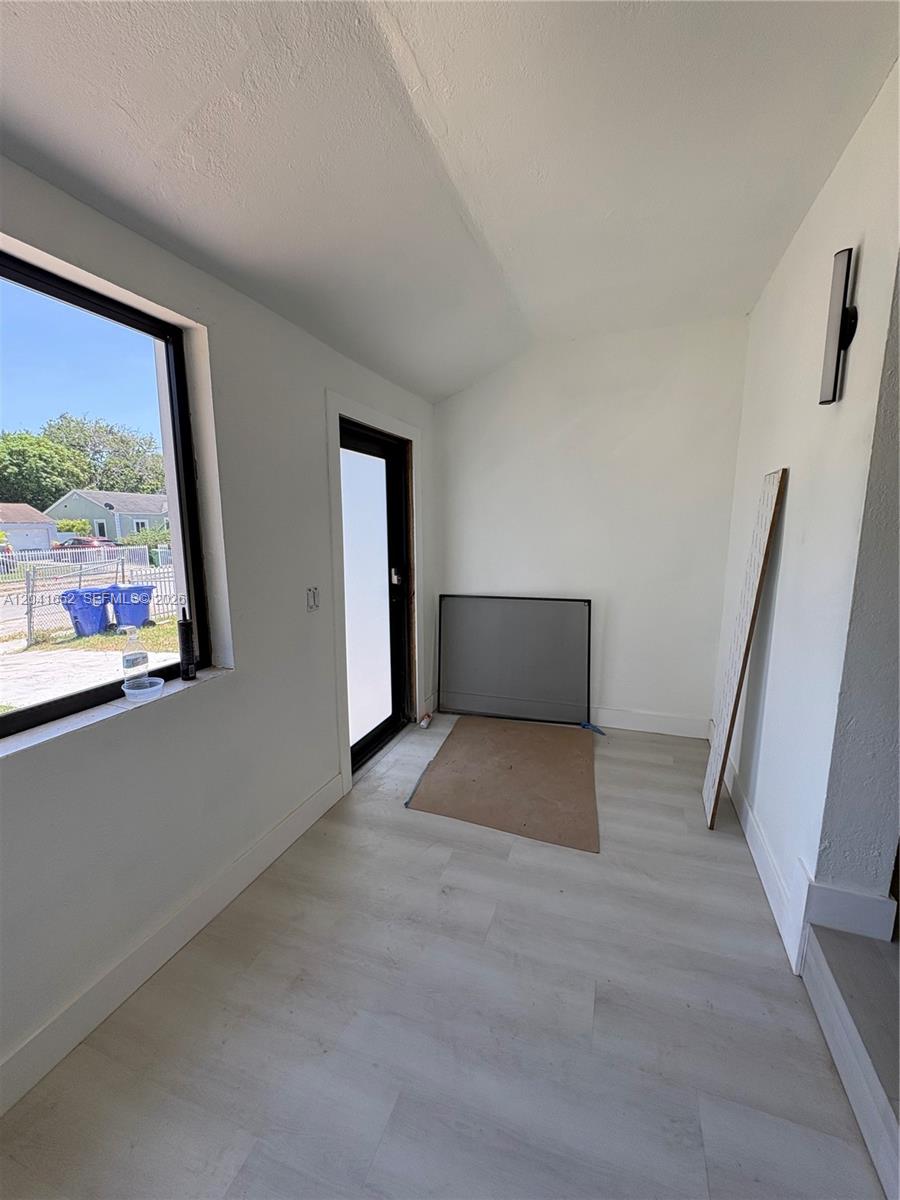 An empty room features white walls, light wood-look flooring, a large window, and a door — 811 NW 65th St, Miami FL 33150, MIAMI, FL