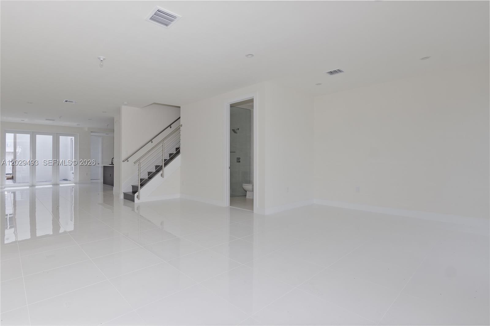 A bright, minimalist living space features white tile floors, a staircase, and an adjacent bathroom — DORAL, FL