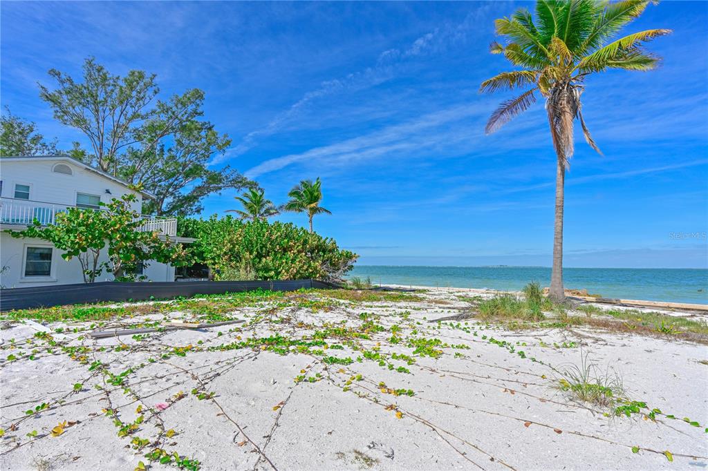 A sandy beach area with low-lying vegetation sits in front of a white house — 837 N SHORE DRIVE, ANNA MARIA, FL