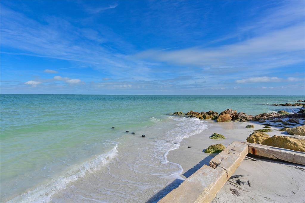 A sandy beach meets turquoise water with rocky shoreline and a concrete structure — 837 N SHORE DRIVE, ANNA MARIA, FL