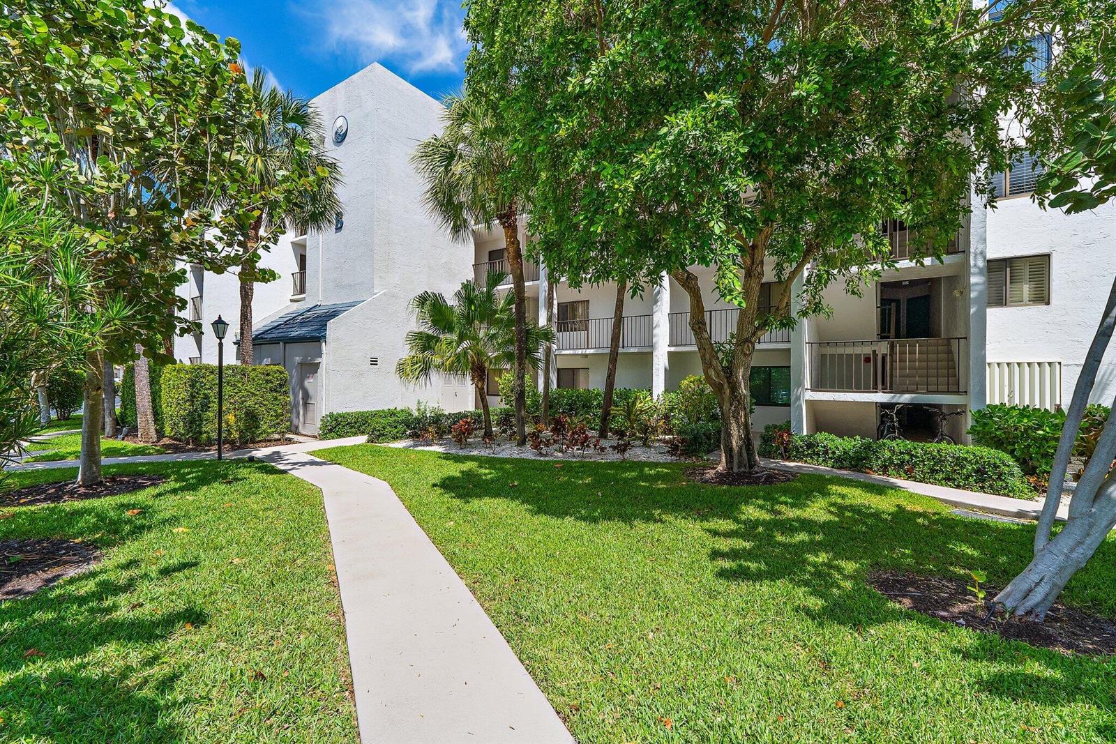 A concrete walkway leads toward a multi-story white apartment building surrounded by trees and grass — 1605 S Us Highway 1 # B205, Jupiter FL 33477, JUPITER, FL