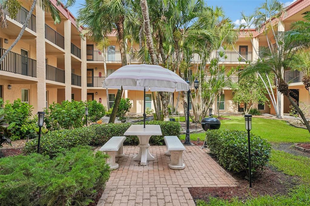 A paved patio with a table, benches, and umbrella sits in a courtyard between apartment buildings — 8911 BLIND PASS ROAD UNIT 104, ST PETERSBURG, FL