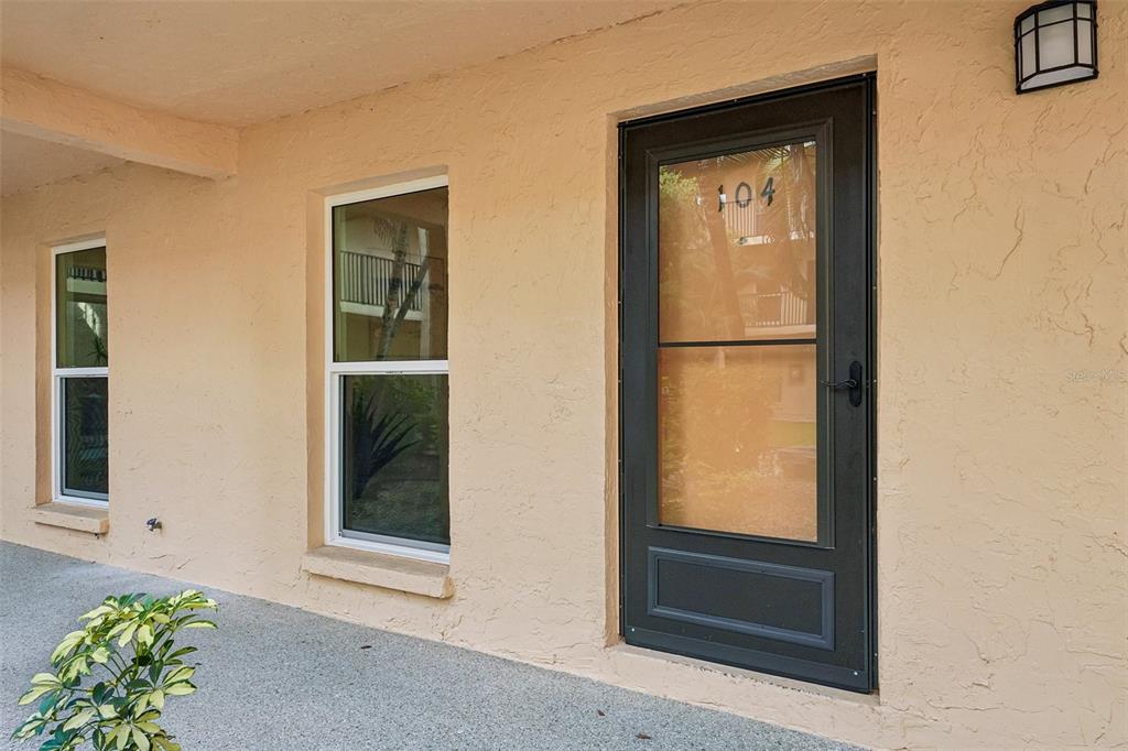 A beige exterior wall features a dark-framed storm door labeled 104 and two adjacent windows — 8911 BLIND PASS ROAD UNIT 104, ST PETERSBURG, FL