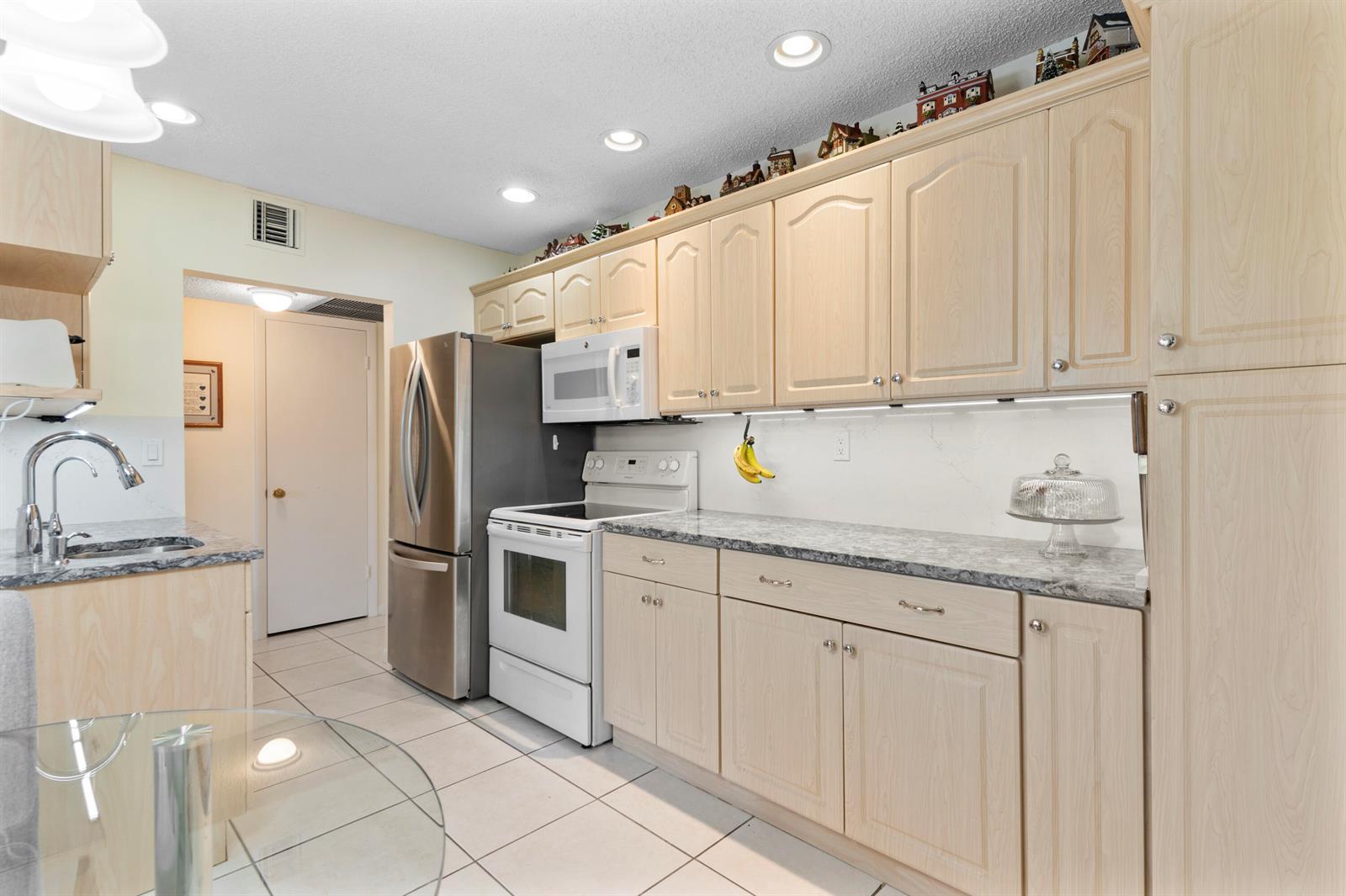 A kitchen features light wood cabinets, granite countertops, stainless steel appliances, and white tile flooring — 5180 E Sabal Palm Boulevard # 234, Tamarac FL 33319, TAMARAC, FL